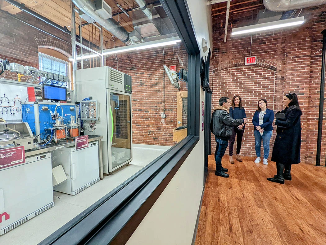 Four people discuss manufacturing beside a glass wall overlooking process equipment and control screens.