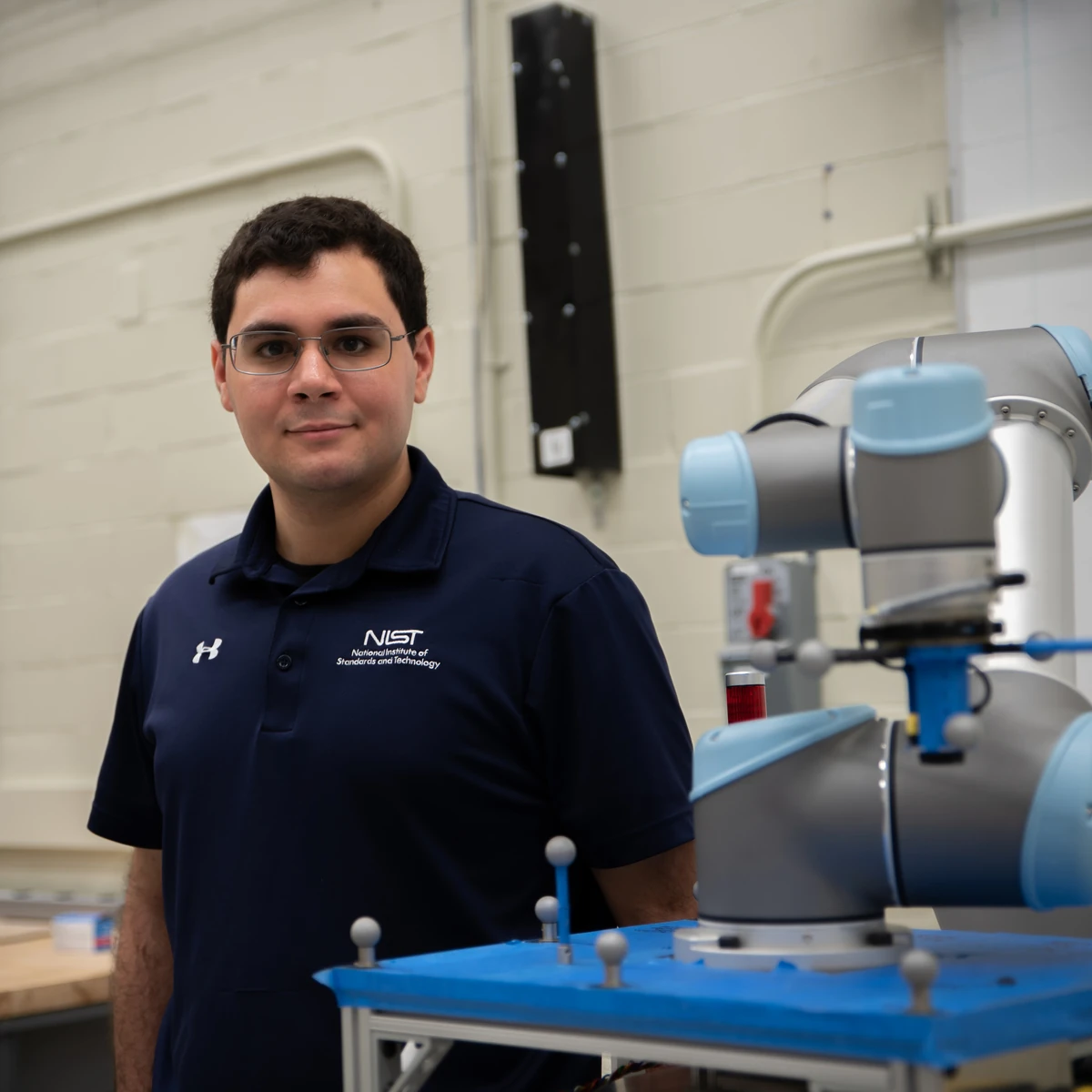 NIST computer scientist Omar Aboul-Enein beside a silver and blue robotic arm and measurement apparatus in a laboratory.