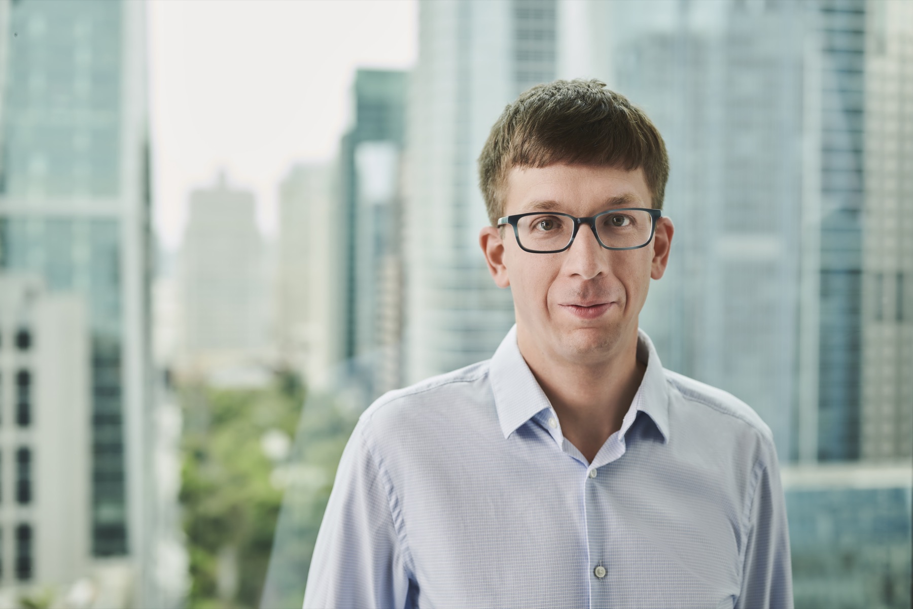 Matei Zaharia wears glasses and a light blue shirt in front of large office windows.
