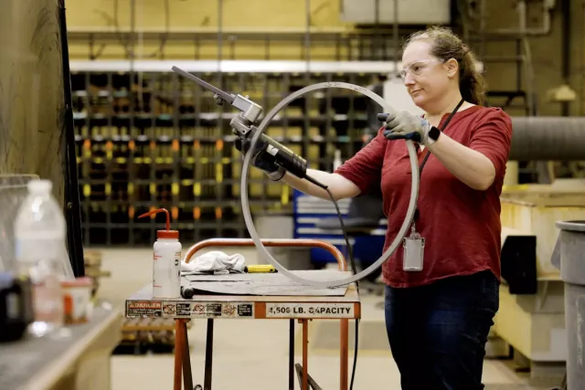 Engineering technician Kristin Lee, wearing safety glasses and gloves, uses a powered tool on a large metal ring in a machine shop
