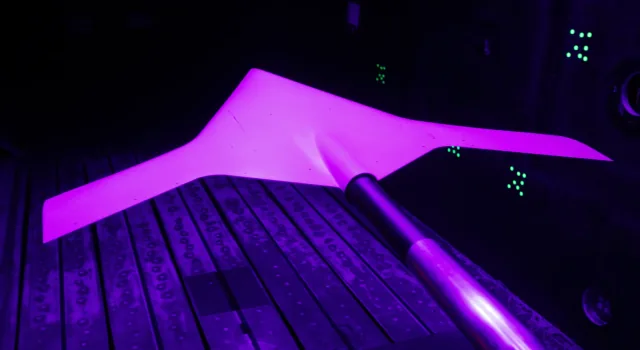 NASA’s Swept Wing Flow Test model glowing pink under ultraviolet light inside a dark wind tunnel.
