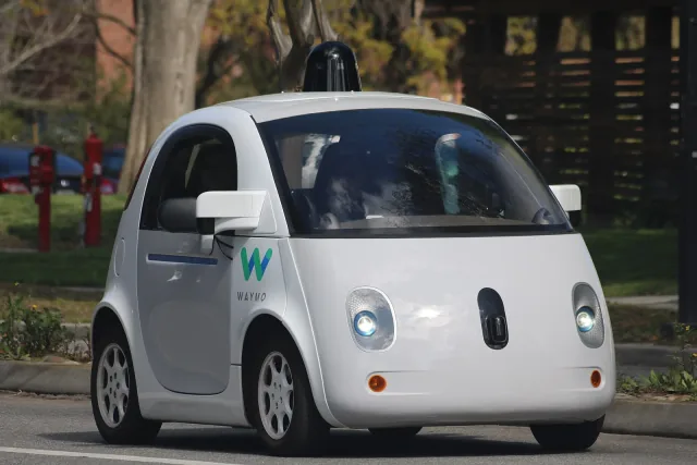 A white Waymo self-driving test car with roof-mounted sensors travels on a road in Mountain View.