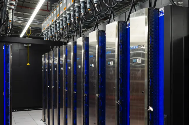 Rows of metal cabinets with blue-lit grilles and overhead cables in the Frontier supercomputer facility.